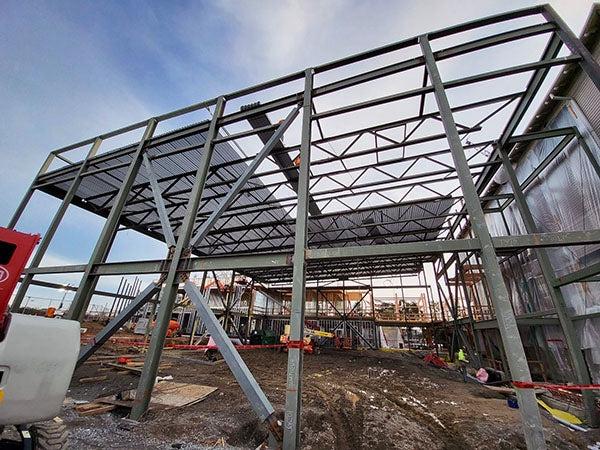 a structural steel frame for a building with an existing building tarped off next to it