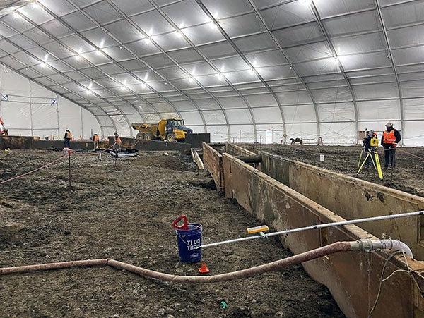 metal forms stand on edge in dirt under a giant tent with lights. construction workers are using equipment