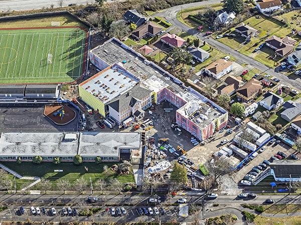 aerial of large building under construction, another large building at the lower right, and a green turf athletic field