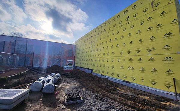 a 2 story solid wall with yellow sheathing adjoins a perpendicular building under construction that is covered in plastic. dirt and construction equipment are in the foreground