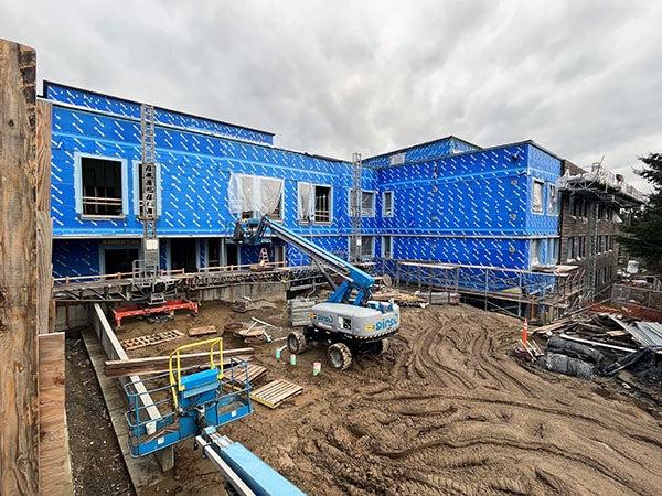 aerial showning a large L shaped building and construction site. the building has blue sheathing and a lift truck