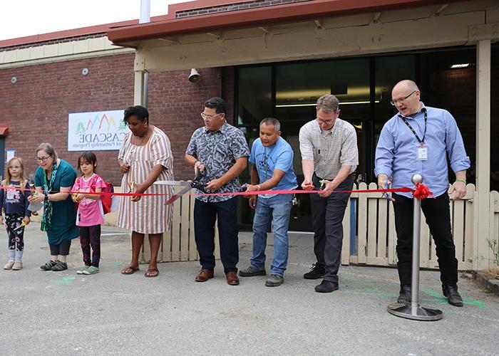 a group of people are lined up behind a red ribbon. all hold scissors with the person in the middle holding giant scissors