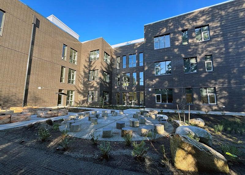 an outdoor area has blocks on a paved area, large rocks, and some plants. A large building with windows is behind it.