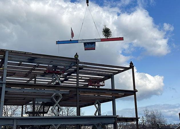 a steel beam is being slung into place at the top of a steel framed building by a crane . Two people wait on top of the existing structure