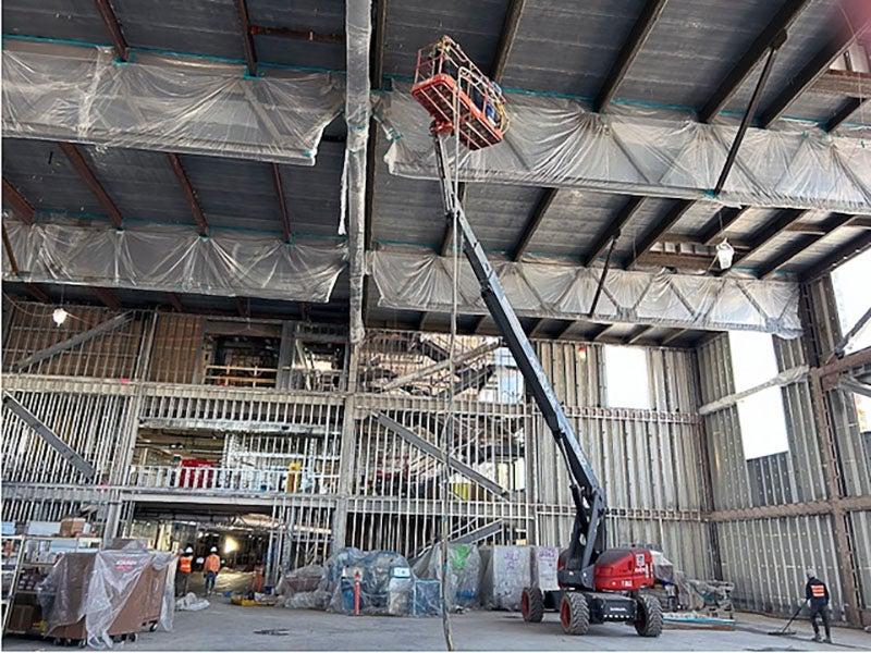 inside a building under construction with metal framing. a lift has a person lifted two the ceiling of the multi-story space