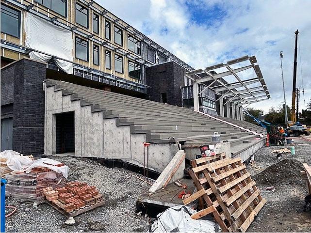 a building under construction with concrete bleachers under construction in front and a frame for a roof over part