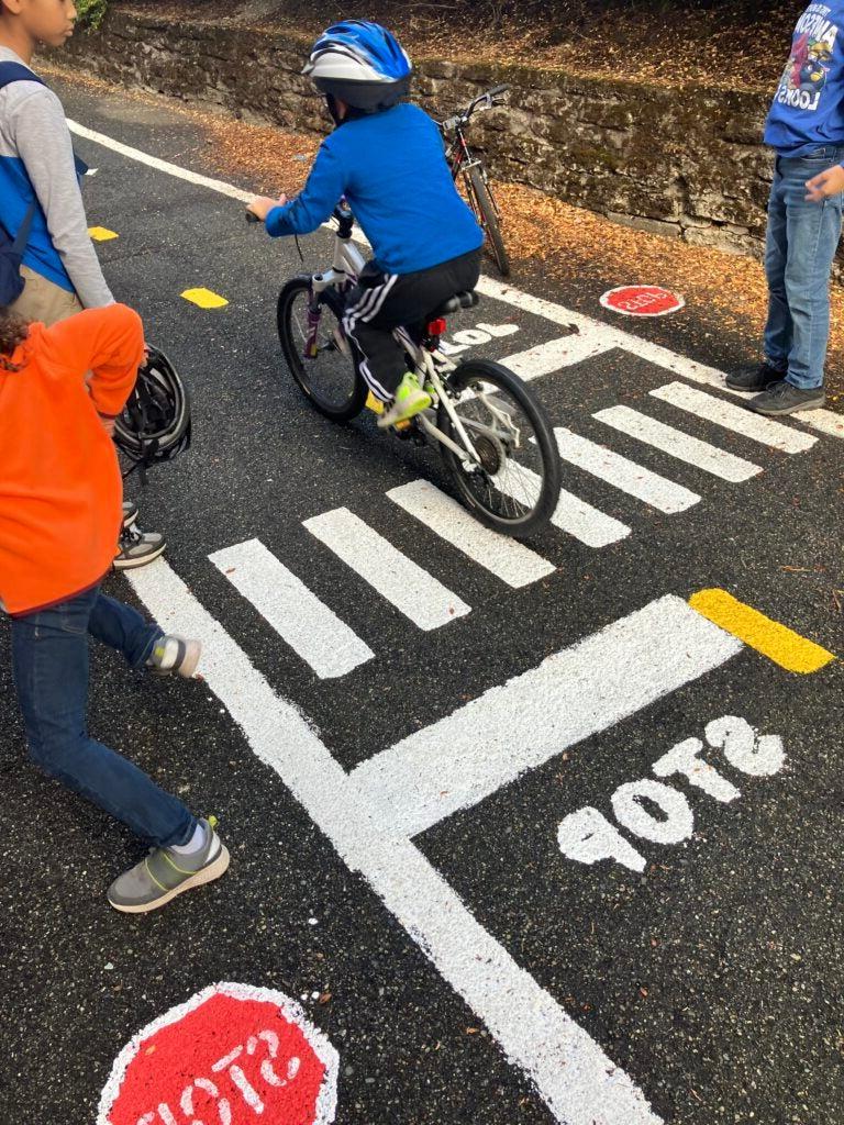 student practices biking on miniature painted street. Other students cheer them on.